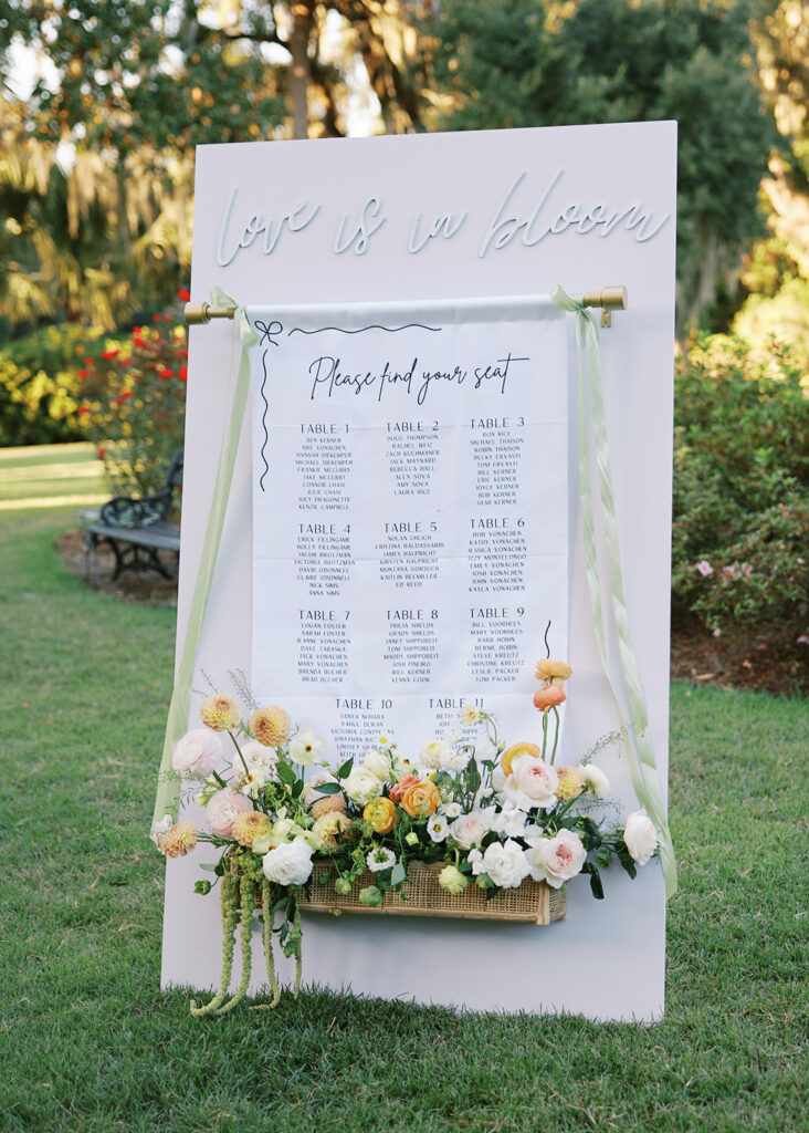 A custom seating chart display featuring raised script that reads "Love is in Bloom" atop a board with a hanging tapestry with the tables and guest names tied with sage green satin ribbon and a rattan flower box at the bottom with lush bright blooms.