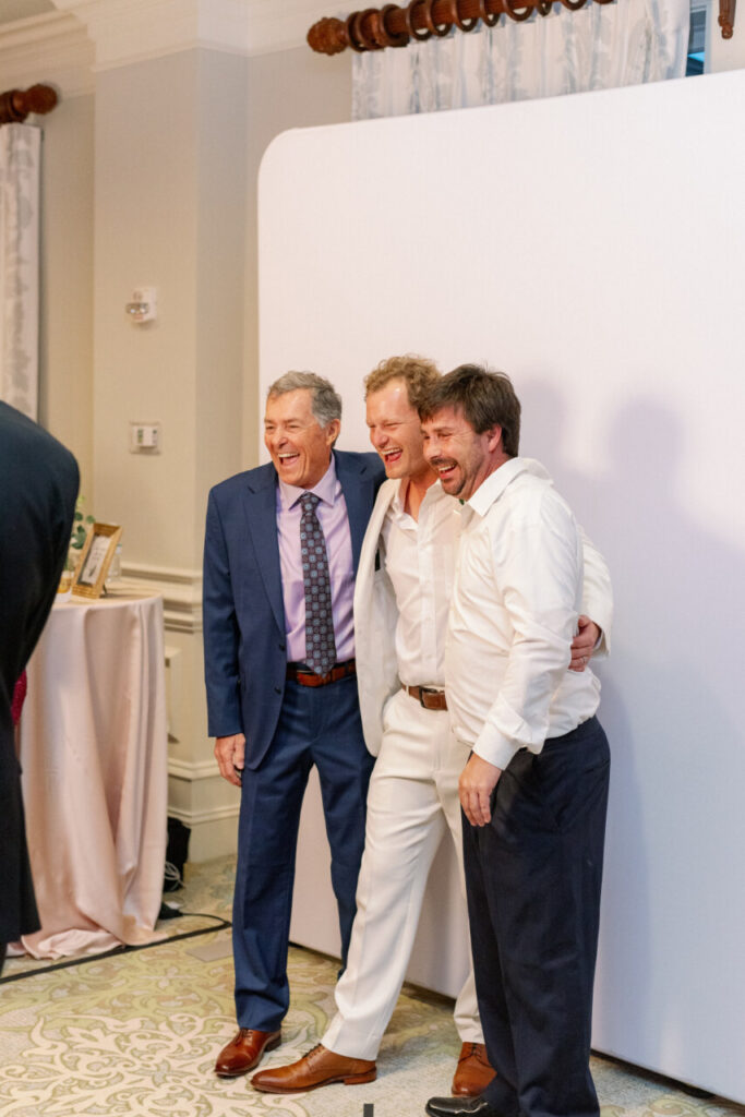 A groom and two wedding guests smile in front of a photobooth at a wedding reception in the ballroom of Seabrook Island Club.