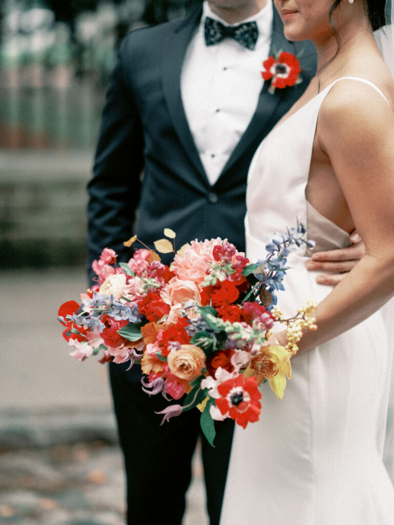 A bride in a white dress with a flower bouquet featuring red, pink, blue, and yellow flowers standing in front of a groom in a black tux with red anemone boutonniere on the lapel.