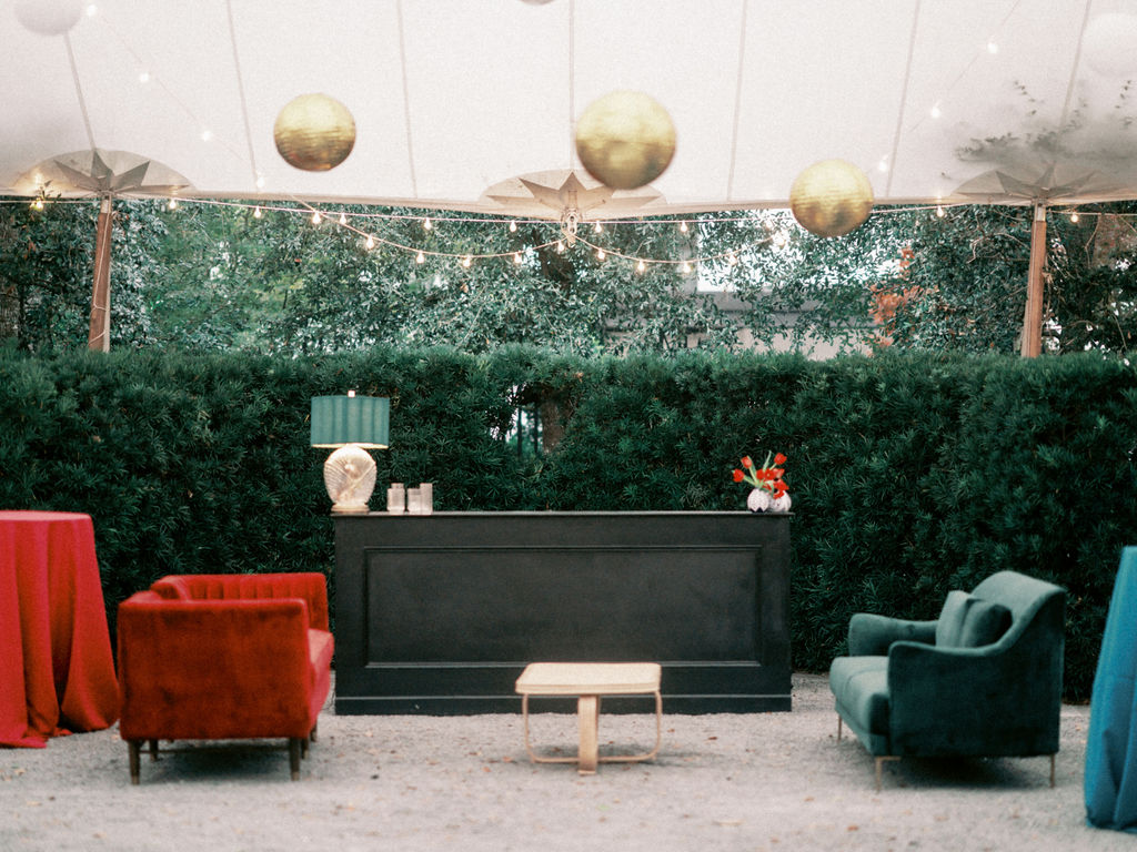 A lounge area at a wedding reception featuring a bright pink velvet couch, emerald green velvet couch, and rattan coffee table in front of a black bar withe red tulip floral arrangements and scalloped emerald green table lamp.