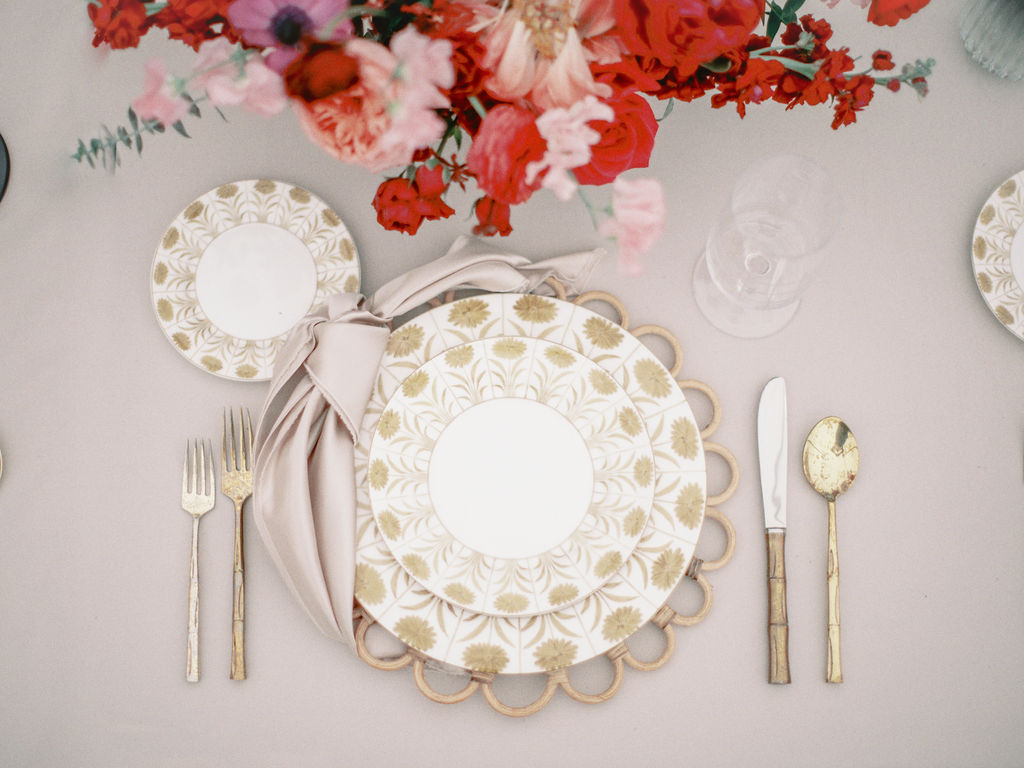 A top down view of a place setting with rattan charger, mustard yellow floral china, and gold metal flatware on top of a taupe tablecloth and red and pink floral arrangement.