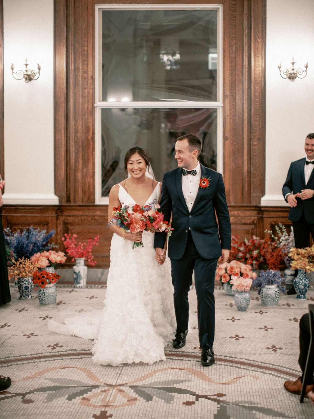 A bride and groom recessing down the aisle after exchanging vows in the rotunda of the Gibbes Museum of Art surrounded by jewel toned monobloom floral arrangments in blue willow chinoiserie urns on the tiled floor.