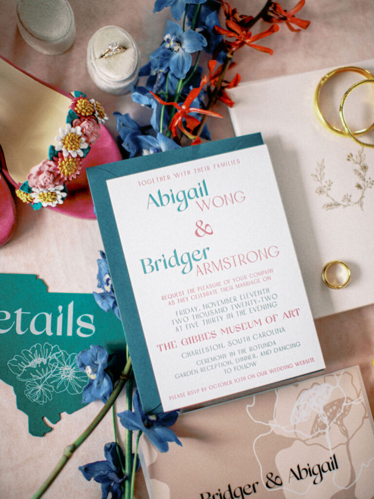 A wedding invitation suite flatway featuring jewel tones in the color palette, custom die-cut details card in the shape of the state of South Carolina, magenta wedding shoes embellished with 3D flowers, an ivory white velvet ring box with rings, and blue and red florals for styling.