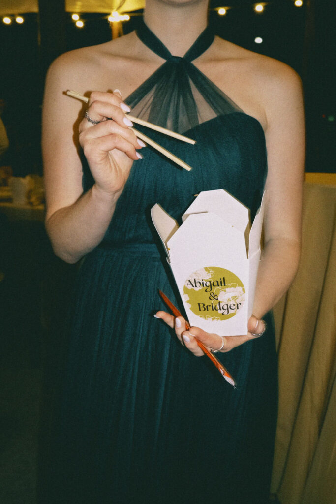 A bridesmaid in a teal dress holding a chopsticks and a Chinese takeout box branded with a custom sticker featuring the couple's names and a signature pattern and font from their stationery suite.