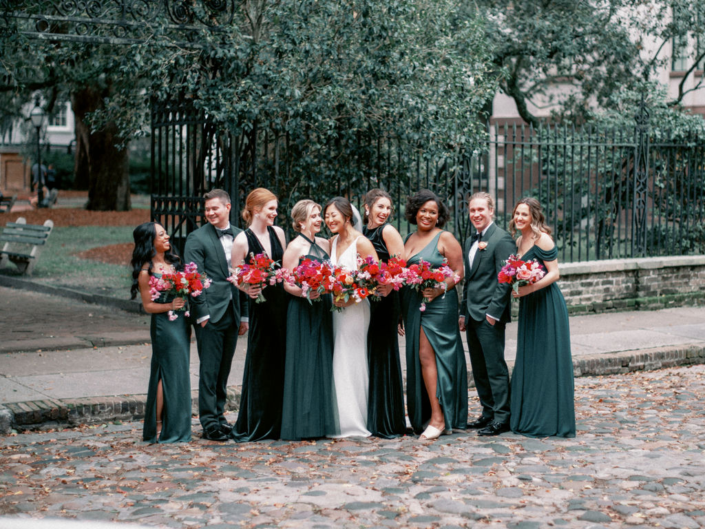 A wedding party wearing emerald green dresses and suits standing next to the bride in a white dress holding red and pink flowers.