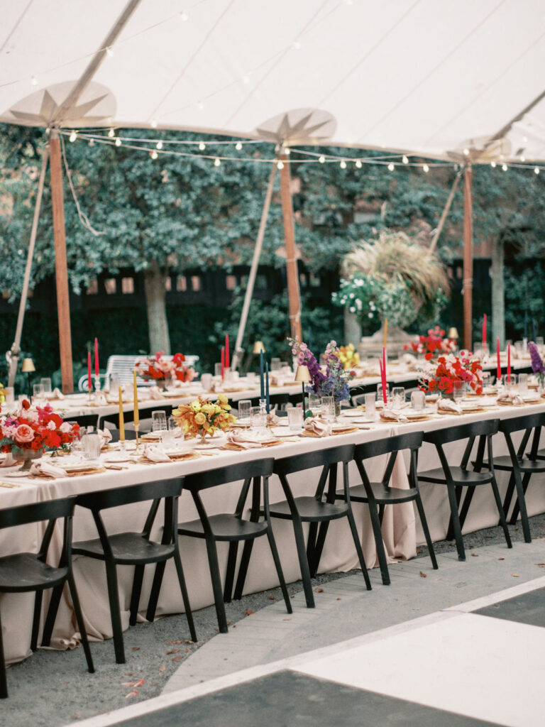 Long reception tables with light taupe tablecloths and black chairs featuring color blocked floral centerpieces in jewel tones of yellow florals, blue and purple florals, and red and pink florals with matching taper candles.