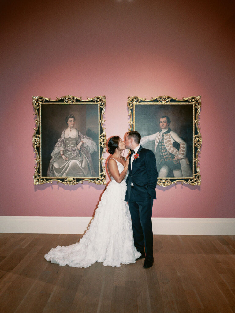 A bride in a white dress and groom in a black tux kissing in front of two oil paintings, portraits of a colonial woman and man in gilded frames hanging on a pink wall at the Gibbes Museum of Art in Charleston, South Carolina.