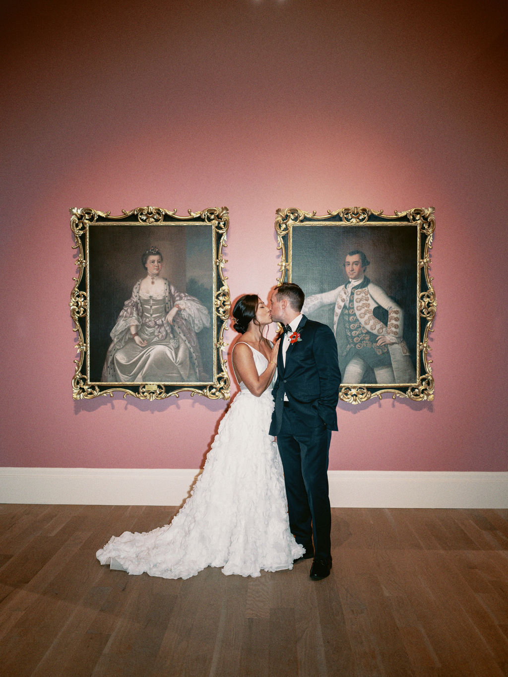 A bride in a white dress and groom in a black tux kissing in front of two oil paintings, portraits of a colonial woman and man in gilded frames hanging on a pink wall at the Gibbes Museum of Art in Charleston, South Carolina.