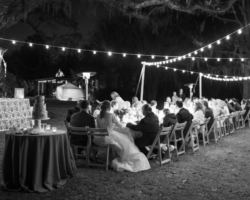 An intimate dinner under string lighting under the oaks with guests seated in wood slatted chairs at a long table with the bride and groom seated at the end with a round table behind them holding a three-tiered wedding cake.