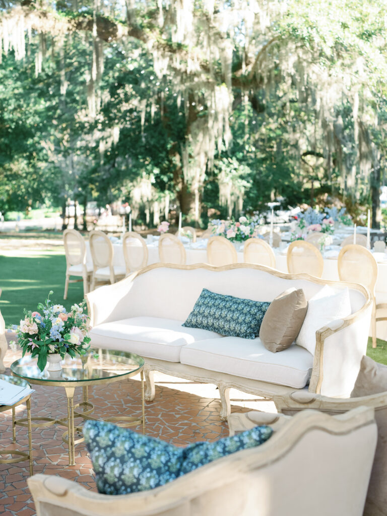 A light beige vintage sofa featuring custom pillows matching the event branding behind a gold and glass-top coffee table with a lush floral arrangement on top.