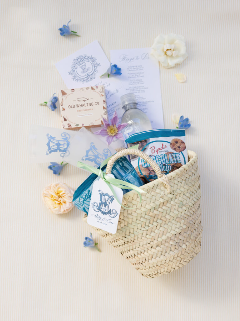 A wedding welcome bag for guests featuring surcies local to the Charleston area: handmade soap bar, Byrd's cookies, Lowcountry Kettle chips, custom monogrammed frosted cups, and a wedding weekend itinerary card packed in a woven basket.