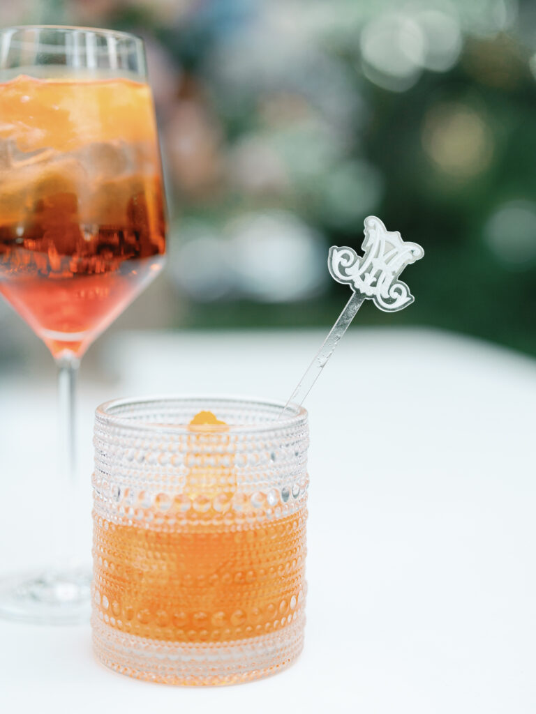 A cocktail in beaded rocks glass with a custom acrylic cocktail stirrer featuring the couple's custom monogram used throughout their wedding branding.