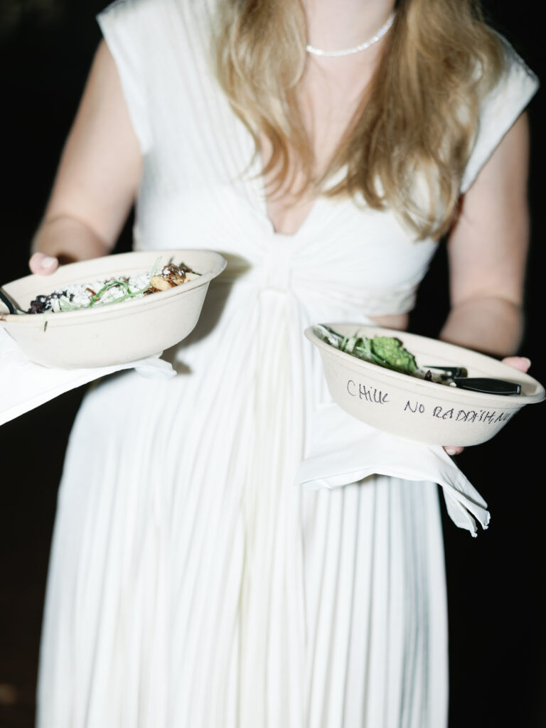 A bride holding two compostable bowls of food at her wedding welcome party at The Lawn of H&I at Riverfront Park in North Charleston, South Carolina.