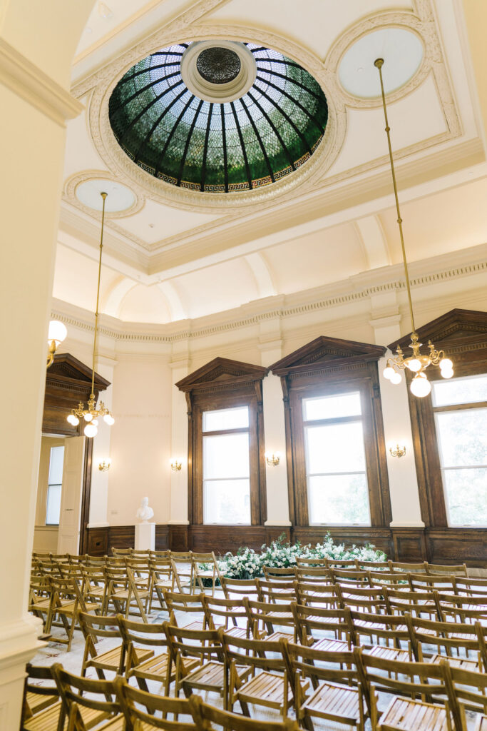 The rotunda of the Gibbes Museum of Art in Charleston, South Carolina set for a wedding ceremony with brown wood slatted chairs and a ground arch at the altar by the windows featuring lush white blooms and greenery.