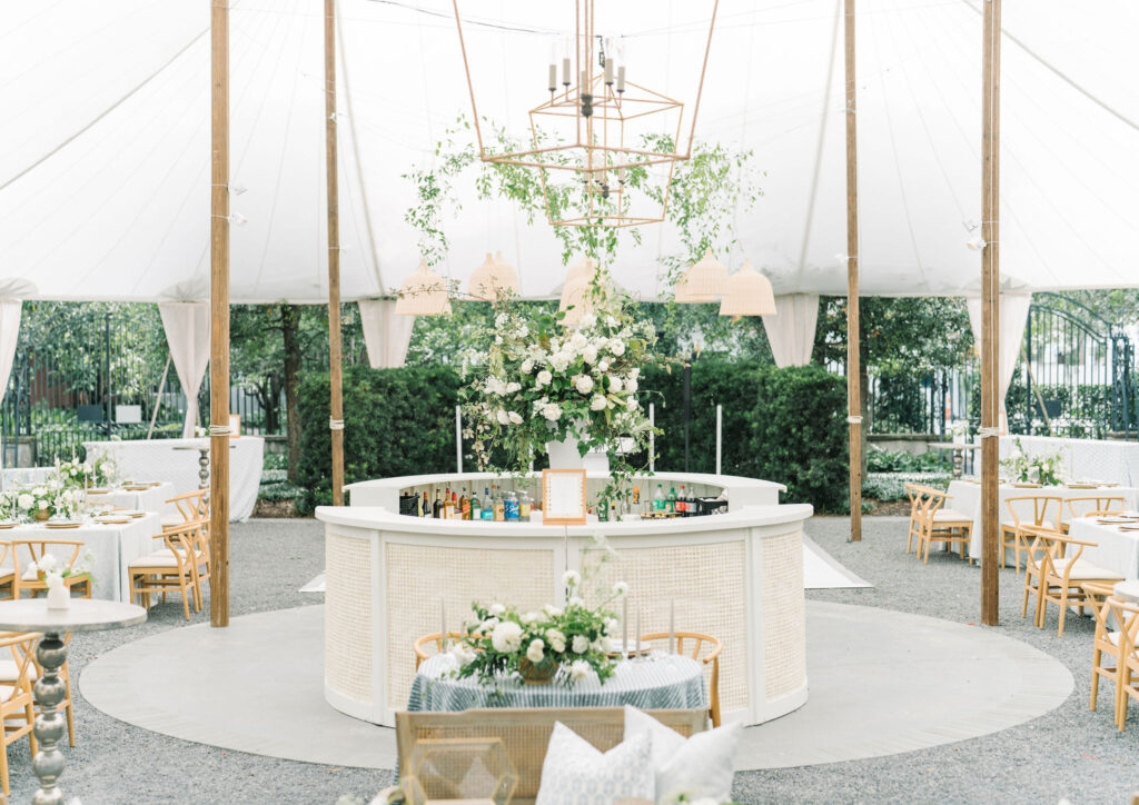 A 360 white bar covered with amate bark paper with a large organically arranged floral centerpiece of white blooms and whimsical greenery in the center under a sailcloth tent in the Lenhardt Garden behind the Gibbes Museum of Art in Charleston, South Carolina.