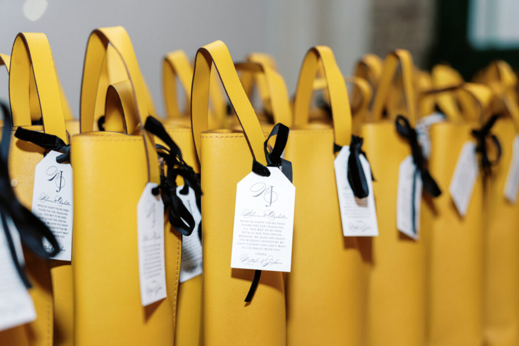 A table filled with custom mustard yellow leather wine totes with itinerary cards featuring the couple's monogram tied with a black velvet ribbon.