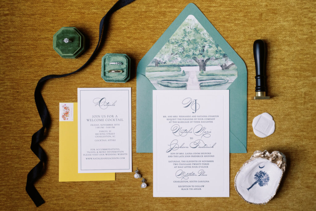 A black letterpress invitation suite flatlay with a watercolor illustration of the Octagonal Garden at Middleton Place on the envelope liner of a sage green envelope and details card on a mustard yellow velvet background with custom monogram wax stamp and green velvet ring box and wedding bands styled with a black velvet ribbon and gold painted oyster shell with South Carolina moon and palm tree mod podged in the inside of the shell.
