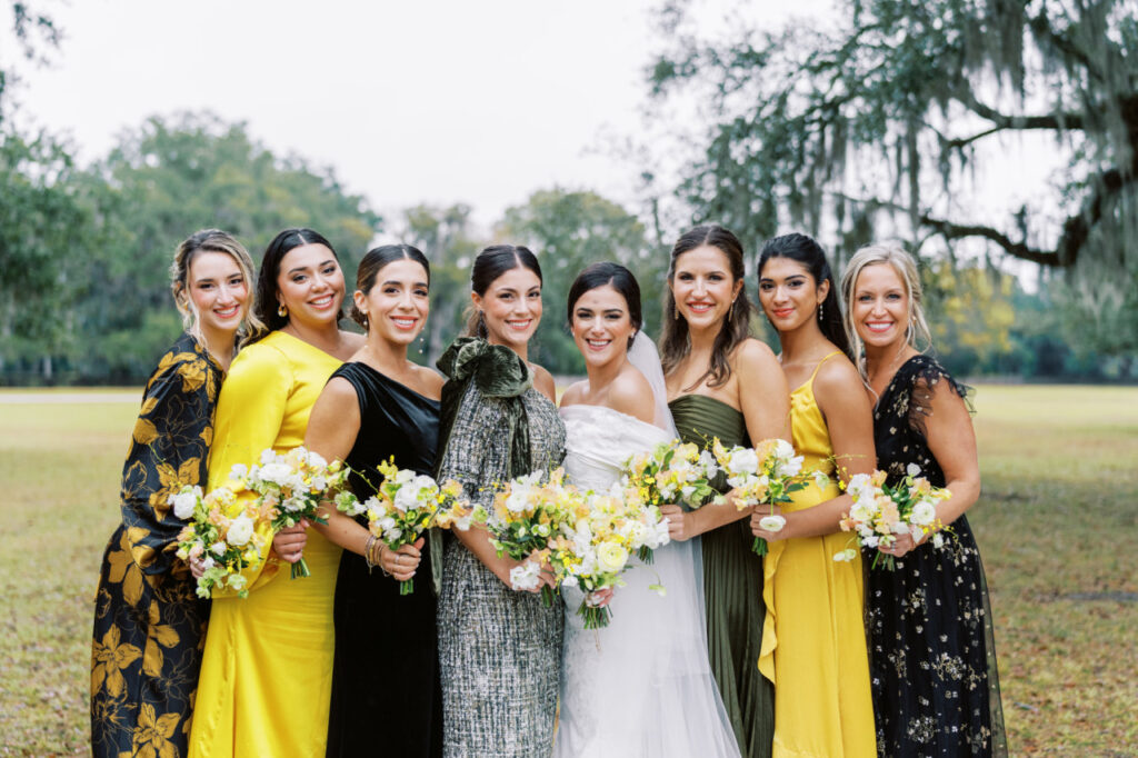 A wedding party portrait featuring bridesmaids in mix-and-matched bridesmaids dresses in different shades, textures, and patterns of yellow, green, and black with a bride in a white dress all holding yellow and white floral bouquets.